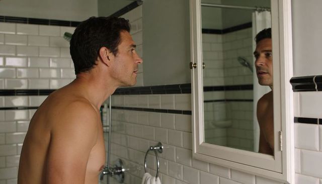 Shirtless man leaning and inspecting reflection in bathroom mirror with subway tile and chrome