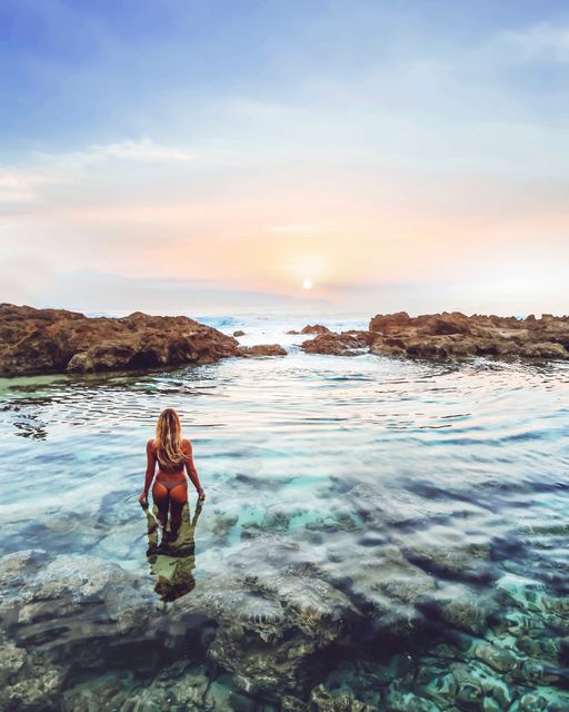 A woman is swimming in a rocky tidal pool as the sun rises. The scene captures the tranquil beauty of nature with the calm water and early morning light. Ideal for depicting relaxation, connection with nature, freedom, and outdoor activity. Suitable for travel advertisements, wellness and lifestyle content, blogs, and inspirational posters.
