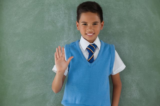 Young boy in school uniform raising hand in classroom, standing in front of chalkboard. Ideal for educational materials, school promotions, academic articles, and websites focusing on children's education and learning environments.