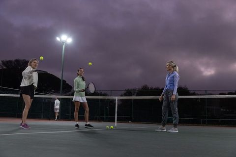 Three women practicing tennis at dusk under floodlights coaching session on outdoor court