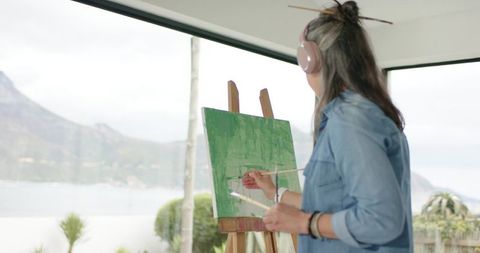 Female artist painting abstract green canvas at easel in coastal studio with ocean view