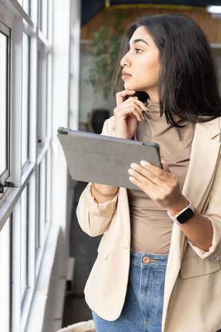 Indian professional woman holding tablet and gazing out window in modern coworking office