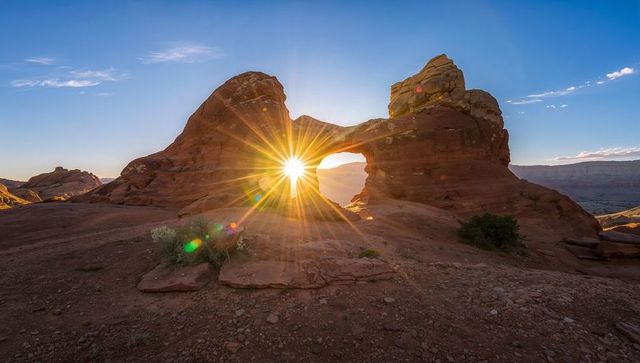 Golden sunburst through natural sandstone arch over desert canyon at sunrise lens flare