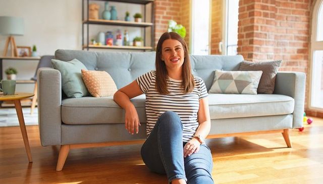 Smiling woman sitting on floor leaning on sofa in sunlit modern living room