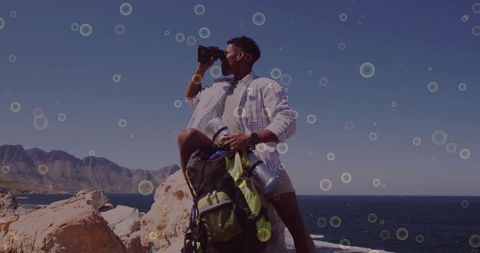 Scanning coastline with binoculars standing on rocky shore with backpack and thermos