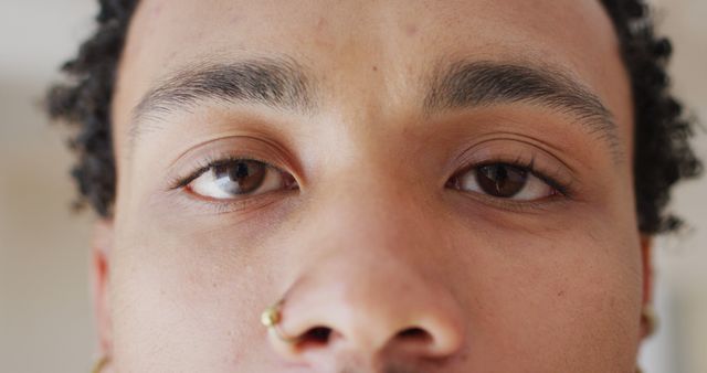 Close-up detail shot highlighting eyes and nose piercing of a young African American male. Can be used in articles or ads focusing on beauty and fashion, personal style statements, or expressing individuality.