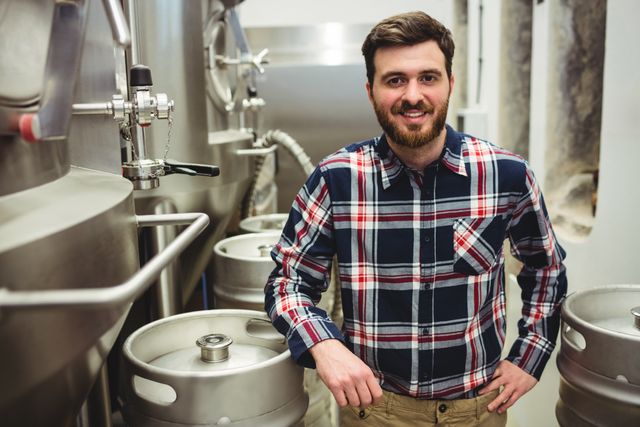 Man standing in brewery next to storage tanks and kegs, smiling confidently. Ideal for use in articles about craft beer production, brewery tours, and the brewing industry. Can also be used in promotional materials for breweries or beer-related events.