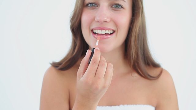 Young brunette woman smiling while applying lip gloss. Great for beauty, skincare, and cosmetic product advertisements or personal care blogs.