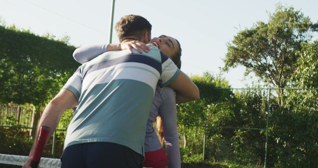 Couple happily embracing on a tennis court after a game, both in casual sportswear and surrounded by lush green trees on a sunny day. Ideal for themes of love, sports, relationships, and outdoor activities. Perfect for use in advertising, blogs, social media posts, or sports and lifestyle magazines.