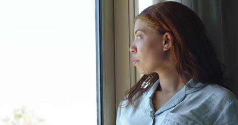 Contemplative woman gazing out window in blue-gray top, side profile, natural light