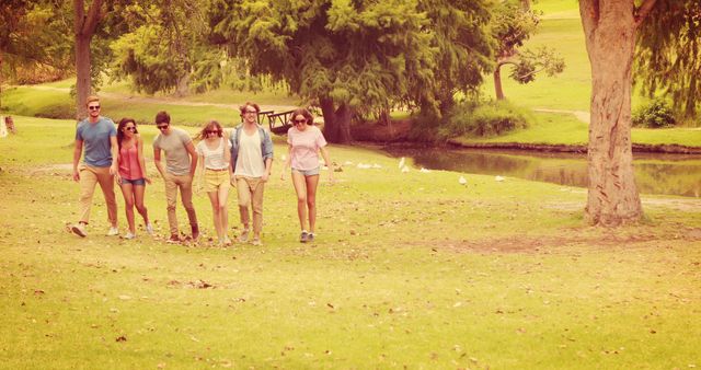 A diverse group of young adults is walking through a park on a sunny day, with copy space. They appear to be enjoying a leisurely stroll by a pond, surrounded by greenery and nature.