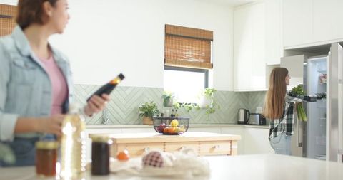 Sunlit modern kitchen two women preparing meals using refrigerator, smartphone and island