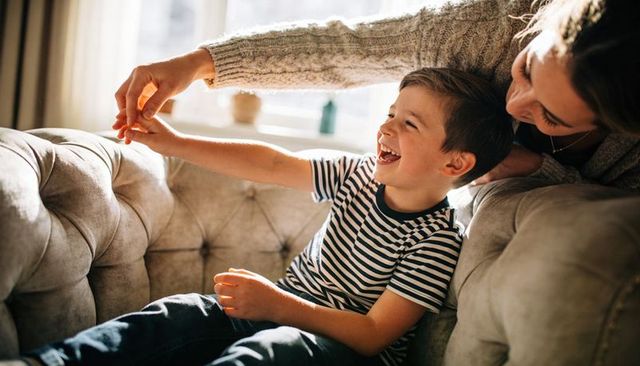Laughing boy reaching toward mother on tufted sofa, sunlit playful cozy family moment