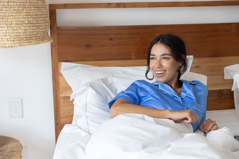 Smiling young woman relaxing in bed wearing blue satin pajamas with wooden headboard