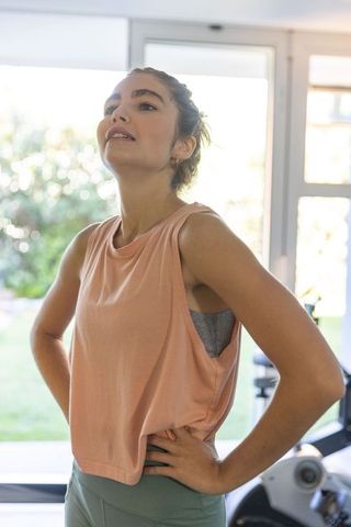 Young woman standing in home gym by glass doors wearing peach workout top and green leggings
