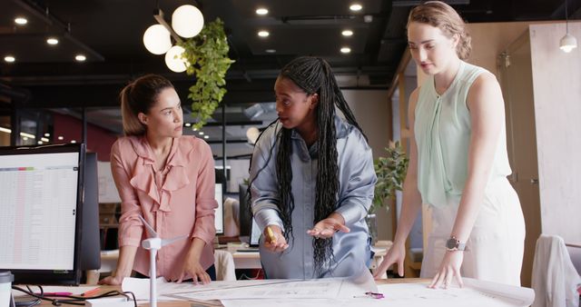 Three female architects engaged in a discussion around large project plans, emphasizing teamwork and collaboration in a modern office. Ideal for use in themes related to workplace diversity, professional collaboration, and creative industries.