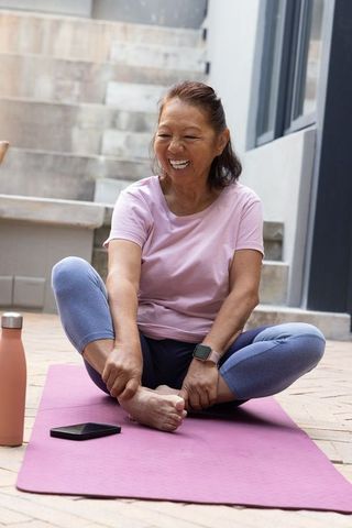 Senior asian woman stretching and smiling on pink yoga mat in courtyard