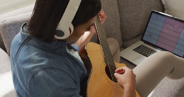 This image shows a woman playing an acoustic guitar while wearing headphones and using online lessons on a laptop. It is ideal for websites or blogs on guitar learning, online music tutorials, home schooling, music practice methods, and lifestyle content.