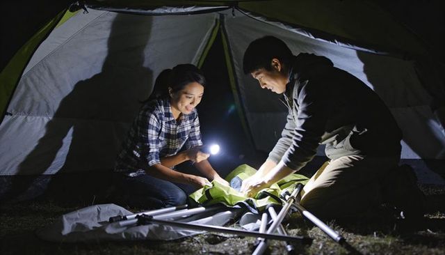 Couple assembling tent at night with flashlight and camping gear, teamwork outdoors