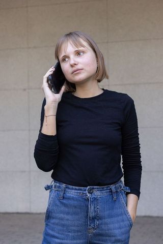 Young woman talking on smartphone against urban tiled wall in black top and denim jeans