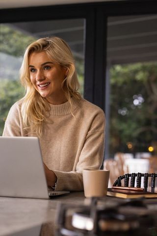 Young woman in twenties working from home at kitchen counter with laptop, mug, notebooks