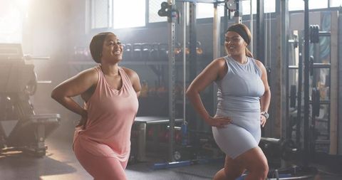 Plus-size black women performing lateral lunges together in gym wearing fitness trackers