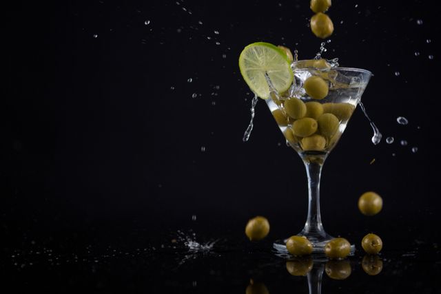 Close-up of olives splashing in to a cocktail martini with lime on table against black background