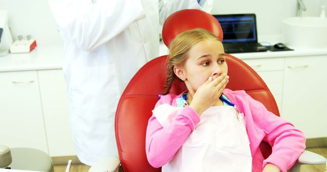 A young Caucasian girl appears nervous while sitting in a dental chair, with a dentist partially visible in the background, with copy space. Her anxious expression and the clinical setting suggest a common fear of dental procedures among children.