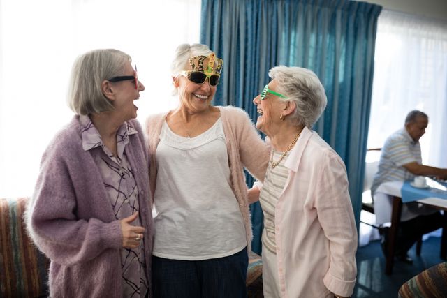 Three senior women are enjoying each other's company while wearing novelty glasses and laughing together in a nursing home. This image can be used for promoting elderly care facilities, highlighting the importance of social interactions among seniors, or showcasing the joy and fun in retirement living.