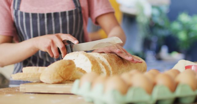 Person wearing apron slicing fresh homemade bread in kitchen. Eggs and other ingredients visible. Ideal for content on baking, culinary skills, home cooking, and fresh ingredients. Suitable for brochures, websites, and cooking blogs l.
