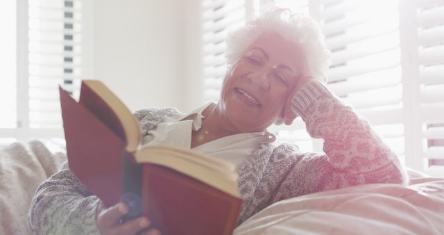 Elderly woman immersed in a good book, reclining comfortably in a bright, sunlit sitting room. Perfect for topics on senior activities, leisure for older adults, promoting relaxation and reading, and aging gracefully.
