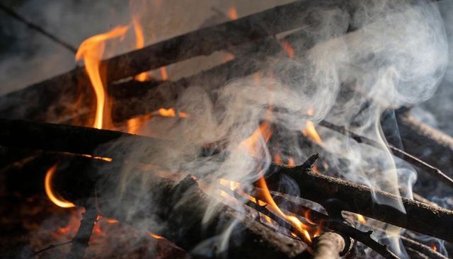 Smoldering campfire logs sending orange flames and gray smoke over charred forest floor