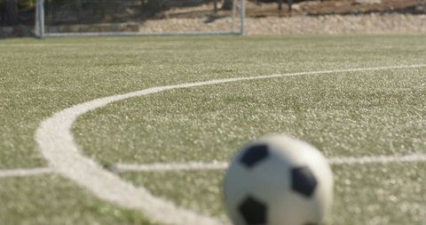 Soccer ball resting on sunlit artificial turf near curved center circle with distant goal