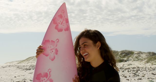 Young biracial woman holds a pink surfboard on the beach. She's ready for a surfing session, showcasing a mix of adventure and relaxation outdoors.