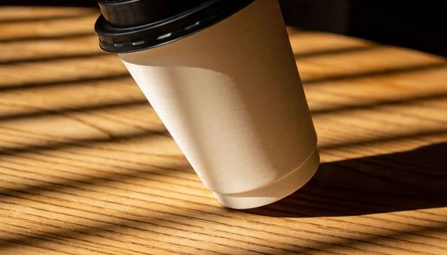 Disposable coffee cup with black lid resting on wooden table casting diagonal sunlight shadows