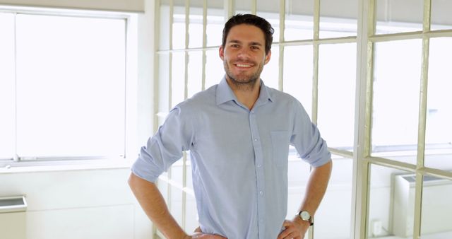 Young man standing with hands on hips in bright office. Perfect for business, corporate, or human resources materials. Great for emphasizing confidence, positivity, and modern corporate culture in marketing and promotional materials.