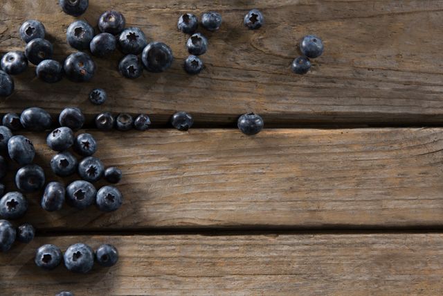 Fresh blueberries scattered on a rustic wooden table. Ideal for use in food blogs, healthy eating promotions, organic product advertisements, and summer harvest themes. The natural wood texture adds a rustic feel, perfect for backgrounds or culinary presentations.