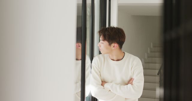 Young man standing indoors and looking out window with arms crossed, appearing thoughtful. Captures a moment of reflection and introspection. Ideal for concepts related to thinking, home environment, contemplation, isolation, and casual lifestyle themes.
