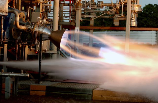 RS-88 rocket engine undergoing a test fire at NASA's Marshall Space Flight Center in Huntsville, Alabama. Powerful engine blast seen against industrial backdrop. Useful for illustrating advanced aerospace testing, rocket engine development, and research at NASA. Ideal for articles or educational materials focused on aerospace engineering, space exploration, or space flight safety systems.
