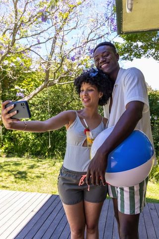 African american couple taking selfie on sunny backyard deck with beach ball and drink