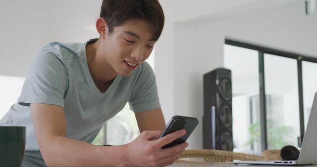 Asian male teenager using smartphone and smiling in living room. spending time alone at home.