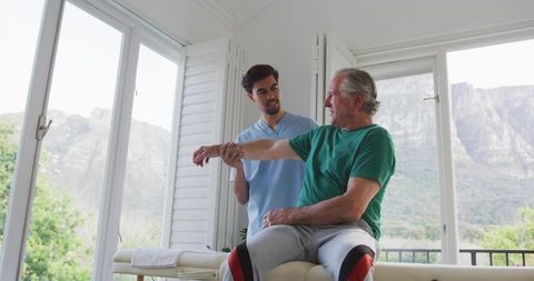 Physiotherapist assisting senior man with shoulder stretch in bright treatment room, mountain view