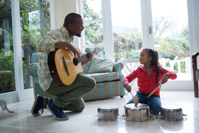 Father and daughter enjoying a creative music session in their living room. The father is playing a guitar while the daughter uses kitchen utensils as drums. This image can be used for themes related to family bonding, creativity, parenting, and home activities. Ideal for articles, advertisements, and social media posts promoting family time, music education, and playful learning.