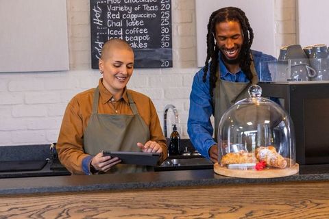 Diverse baristas smiling while using tablet and arranging pastries behind counter