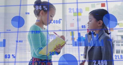 Two girls collaborating on data writing notes on yellow notebook with blue charts overlay