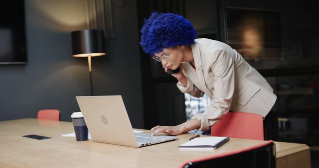 Businesswoman with blue afro hair multitasking by talking on the phone and working on her laptop in a modern office. Good for illustrating themes of diversity in the workplace, multitasking, business communications, professional work environments, and technology use.
