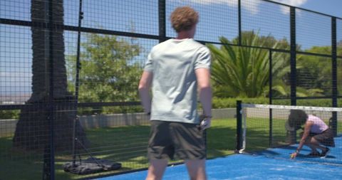 Casual padel duo on blue outdoor court, man holding paddle and partner retrieving ball