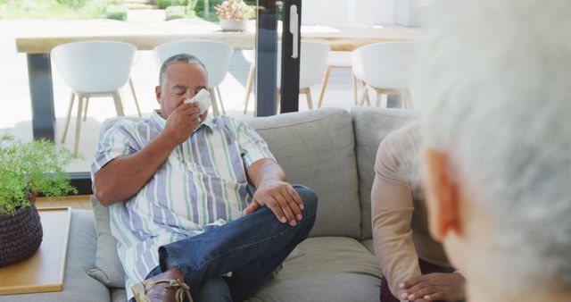 Man having an allergy attack while sitting on a comfortable sofa in living room. Use for topics related to health, allergies, cold and flu season, home comfort, and indoor activities.
