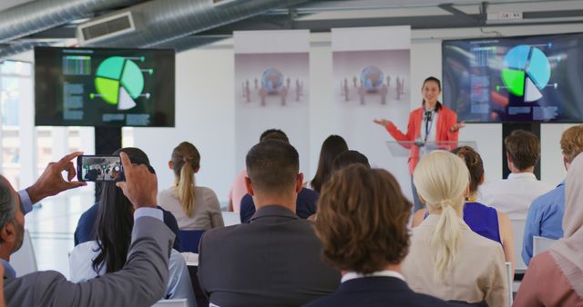 Businesswoman presenting colorful charts and graphs on multiple screens to a seminar audience. Photo suitable for use in business training materials, corporate meetings, professional presentations, or educational seminars.