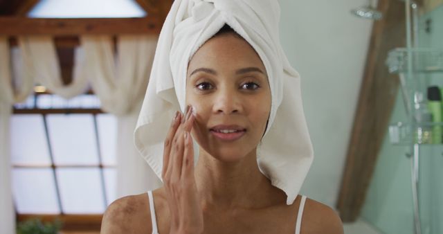 Portrait of biracial woman wearing towel on head applying cream on her face. domestic life, spending quality free time relaxing at home.